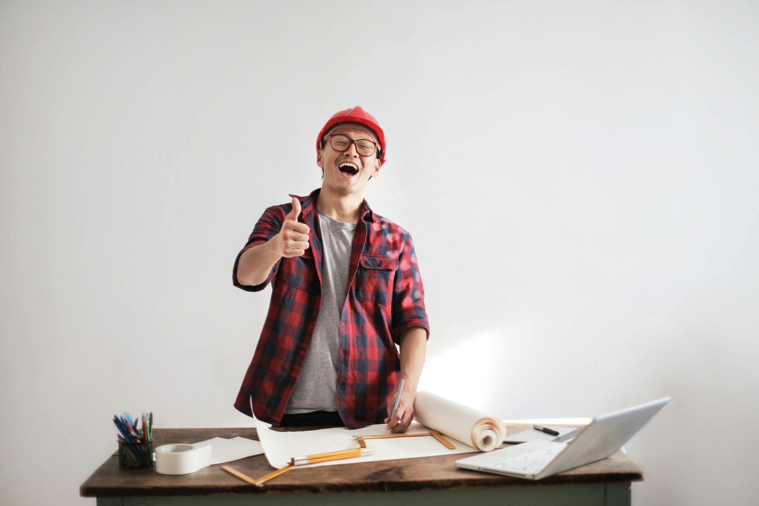 Cheerful male architect in a red hardhat and plaid shirt giving thumbs up at his desk.